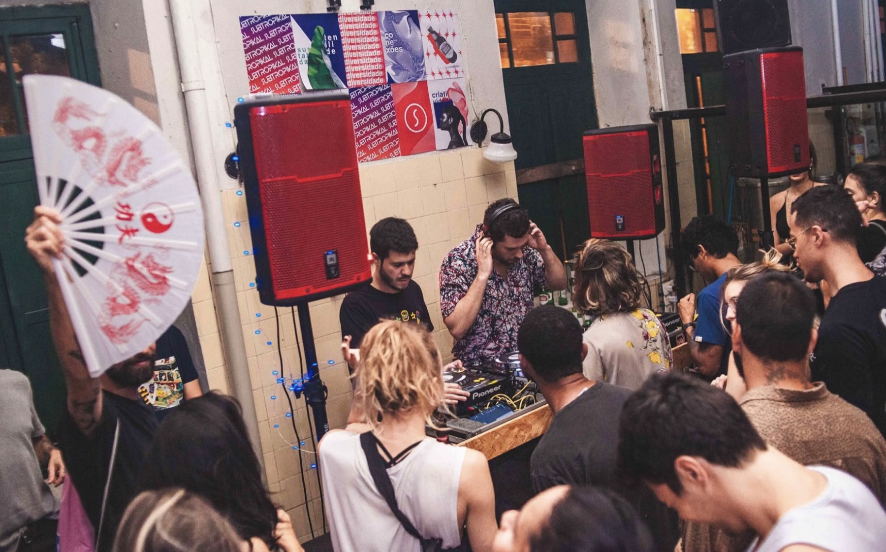 DJ tocando cercado de público num espaço intimista, com cartazes do Subtropikal na parede