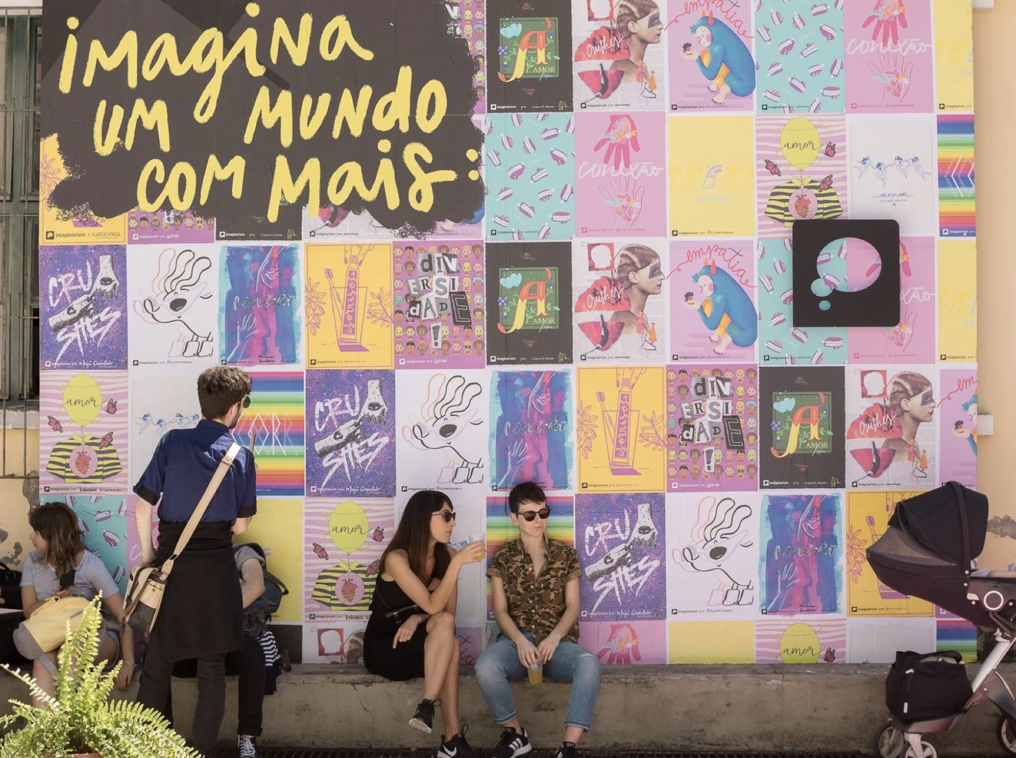 Parede coberta de cartazes coloridos com o mote 'imagina um mundo com mais', colaboração Imaginarium e Subtropikal, com público sentado em frente