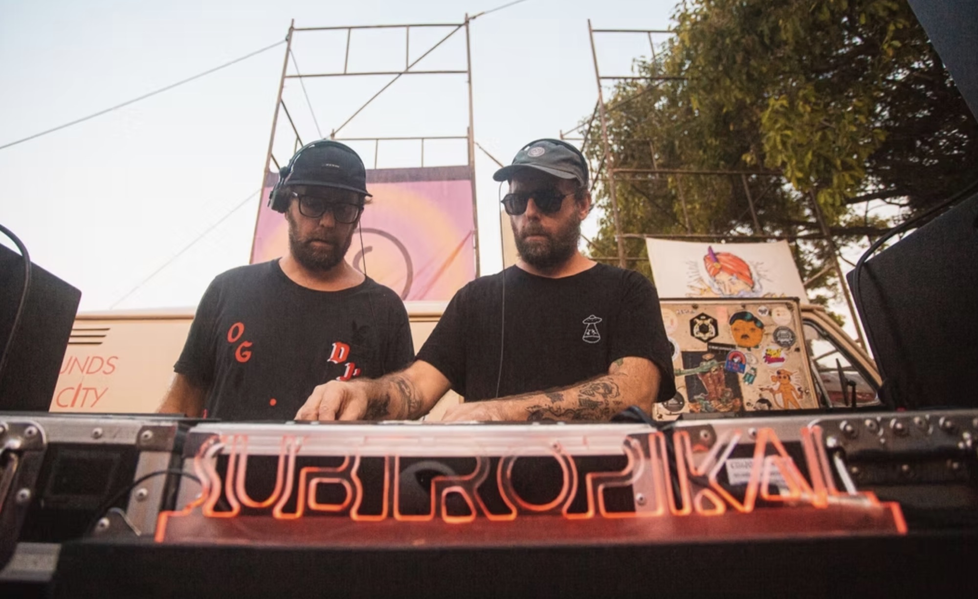 Dupla d'OSGEMEOS em DJ set no fim de tarde, atrás do letreiro em neon do Subtropikal