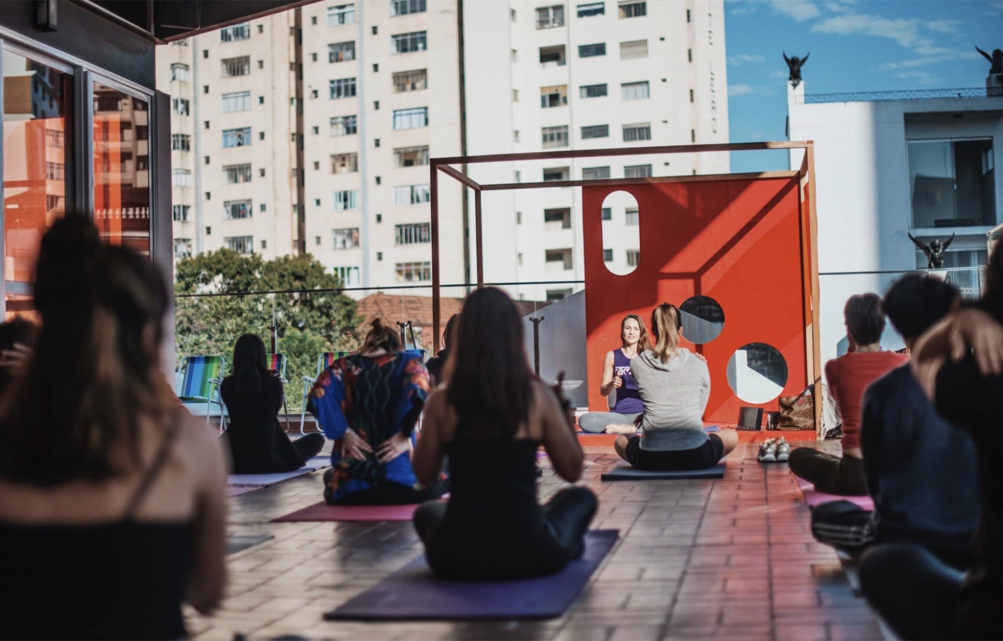 Prática de yoga no rooftop do festival, com público sentado diante de uma instalação vermelha e a cidade ao fundo